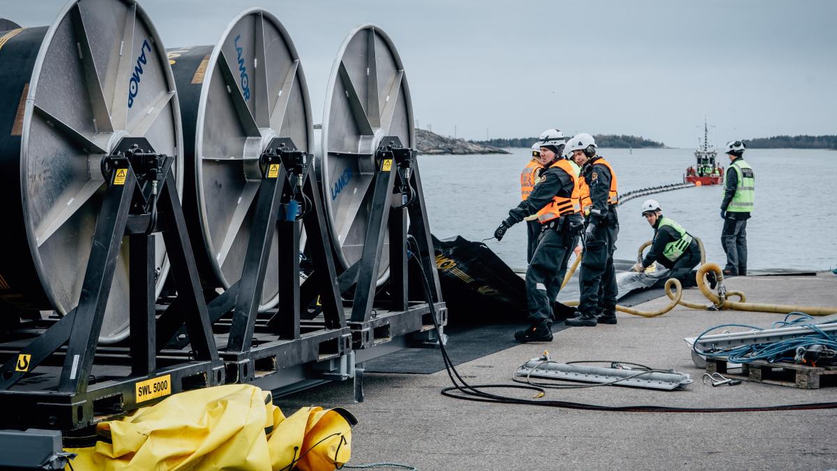 Oil spill response personnel working in a port area with large cable reels and response equipment by the sea.